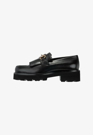 Zwarte leren loafers met dikke gegroefde zool, franjes en een gouden gesp op een witte achtergrond.