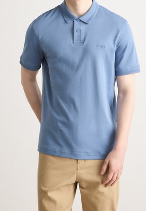 Mann trägt hellblaues kurzärmeliges Poloshirt mit Kragen und beige Hose vor einfarbigem Hintergrund.