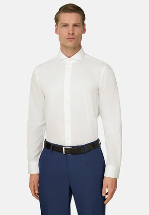 Mężczyzna ubrany w dopasowaną białą koszulę, niebieskie spodnie i czarny pasek, stojący na tle gładkiego białego tła.