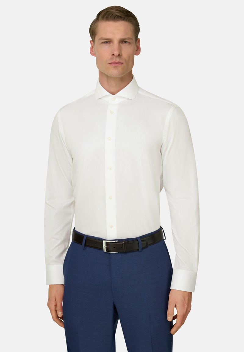 Mężczyzna ubrany w dopasowaną białą koszulę, niebieskie spodnie i czarny pasek, stojący na tle gładkiego białego tła.