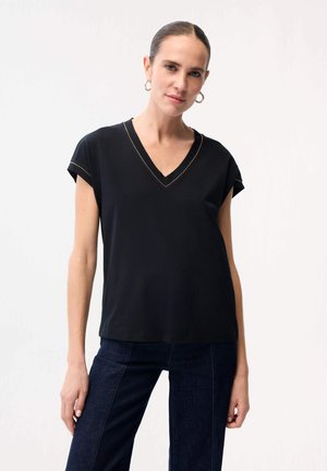 Nainen, jolla on tumma hiukset, musta lyhythihainen V-kaula-aukollinen paita, jossa on kultaiset ompeleet, ja tummat farkut, vaaleaa taustaa vasten.