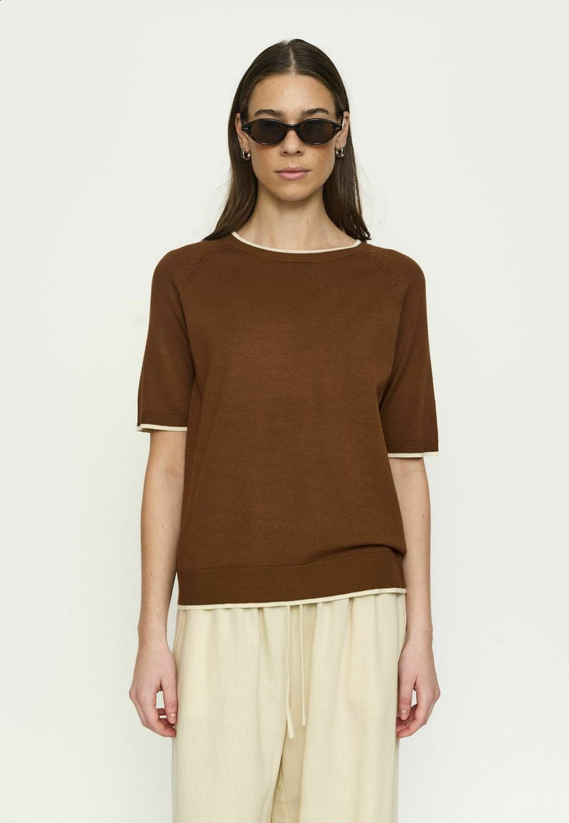 Femme portant un pull à manches courtes marron avec bordure blanche, un pantalon crème à cordon de serrage et des lunettes de soleil fines noires, debout devant un fond clair uni.