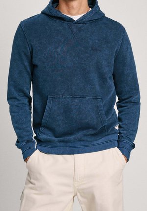 Mann trägt einen ausgewaschenen blauen Hoodie mit Fronttasche und cremefarbene Hose, Hände in den Taschen, steht vor einem einfarbigen Hintergrund.