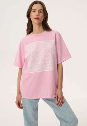 Kobieta z długimi brązowymi włosami nosi luźny różowy T-shirt z białym, skomplikowanym wzorem paisley w kształcie kwadratu oraz jasnoniebieskie dżinsy.