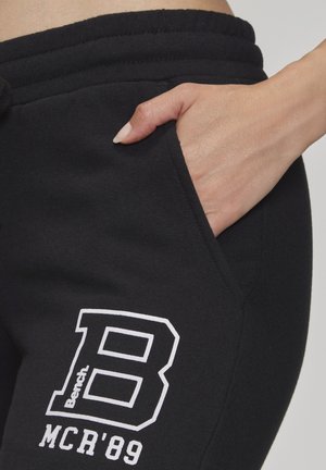Hand resting in black sweatpants pocket with white printed "B Bench MCR '89" logo on upper thigh.