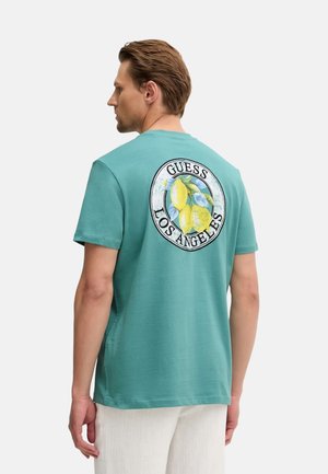 Homme portant un t-shirt bleu sarcelle avec un motif circulaire jaune citron et le texte « GUESS LOS ANGELES » dans le dos, associé à un pantalon blanc.