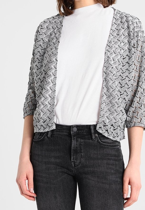 Femme portant un t-shirt blanc sous un cardigan ouvert à motif noir et blanc avec des manches trois-quarts et un jean noir.