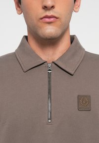 Mann med en taupe zip-up skjorte med krage og et lappemerk design med en ørn og tekst på brystet.