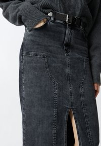 Tamni traper suknja s prednjim prorezom, dva velika bočna džepa i visokim strukom, u kombinaciji s crnim remenom i teksturiranom sivom duksericom.