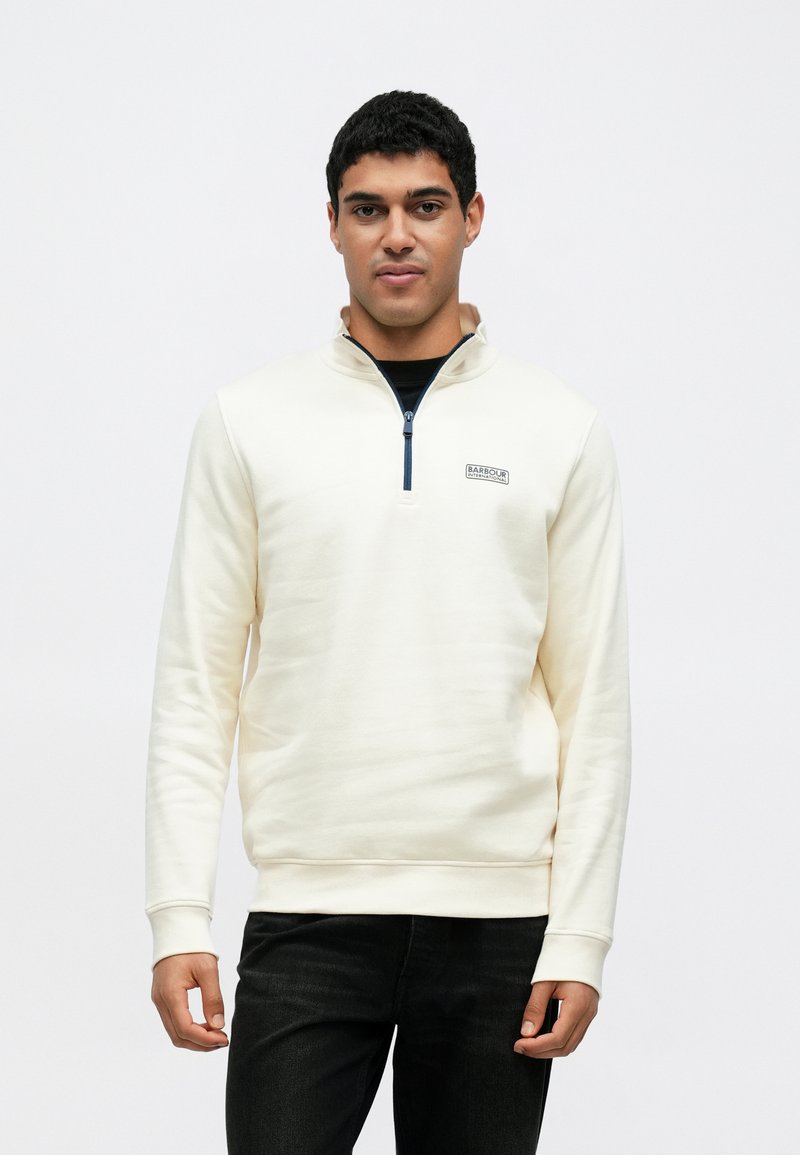 Krém színű cipzáras pulóver magas gallérral, bordázott mandzsettákkal és egy logóval az mellkason, sötét farmerrel párosítva. Simogató tapintású anyag.