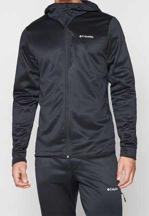 Mann trägt eine schwarze Columbia Zip-up-Kapuzenjacke und passende schwarze Hose mit Columbia-Logos auf Brust und Oberschenkel.