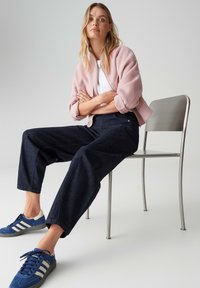 Veste bomber rose clair, t-shirt blanc, jean large bleu foncé, et baskets bleues avec des accents blancs, assis sur une chaise argentée devant un fond gris.