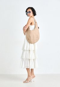 Beige semsket hobo-veske med en buet form, med sølv øyelets langs toppen, båret over en lagdelt hvit kjole med blonder.