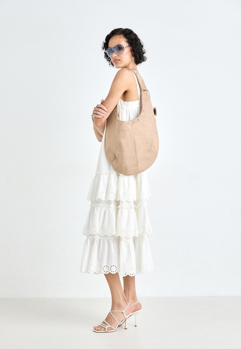 Bolso hobo de ante beige con forma curva, adornado con ojales plateados en la parte superior, combinado con un vestido blanco de capas con acentos de encaje.