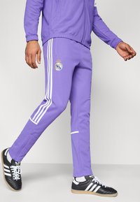 Μωβ αθλητικό παντελόνι με λευκές ρίγες, διακοσμημένο με λογότυπο συλλόγου. Φοριέται με μαύρα και λευκά αθλητικά παπούτσια· το ύφασμα φαίνεται λείο και εφαρμοστό.