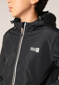 Veste noire imperméable avec fermeture éclair, capuche et accent beige. Présente un patch logo sur la poitrine et une texture lisse.