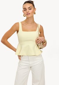 Top peplum en tissu jaune doux, avec un décolleté carré et de larges bretelles, associé à un pantalon large blanc et un sac beige.