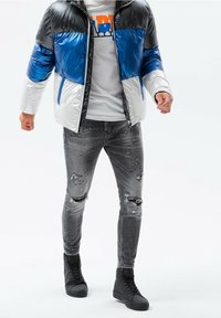 Bunte Steppjacke in Schwarz, Blau und Weiß; graue zerrissene Jeans; schwarze High-Top-Sneaker; getragenes Design und glänzende Textur.