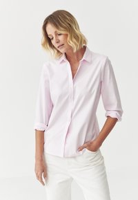 Chemise boutonnée rose clair avec un col évasé, manches longues retroussées et une poche poitrine, associée à un pantalon blanc. Texture en coton lisse.