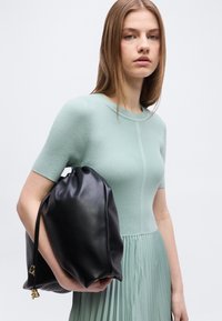 Νεαρή γυναίκα με φόρεμα σε ριμπ ύφανση χρώματος μέντας, κρατώντας μια μεγάλη μαύρη δερμάτινη τσάντα που κλείνει με κορδόνι, σε απλό λευκό φόντο.