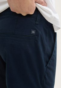 Pantalons bleu marine foncé avec une texture lisse, dotés d'une poche arrière et d'une petite étiquette logo noire. Coupe slim, avec un design sobre et des lignes épurées.