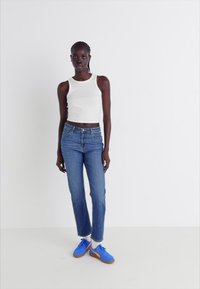 Débardeur court blanc, jeans bleus à jambe droite et baskets bleues avec semelles brunes. Design simple et coupe classique.