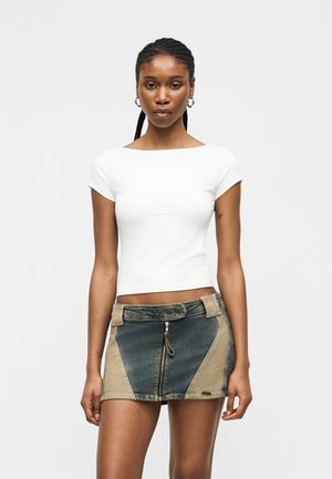 Kvinde med flettet hår iført øreringe i hoop-design, en hvid tætsiddende kortærmet top og et tofarvet denim mininederdel med lynlås foran.