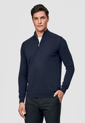 Mężczyzna ubrany w granatowy sweter z długim rękawem i półzamkiem oraz czarne spodnie, stojący z jedną ręką w kieszeni na jednolitym tle.