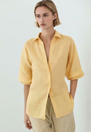 Kvinde med kort blond hår iført en løs, knaplukket gul hørskjorte med opkørte ærmer og beige bukser mod en ensfarvet baggrund.