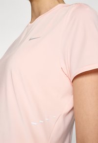 Nike svijetlo ružičasta atletska majica kratkih rukava, s srebrnim logotipom i suptilnim bijelim linijama sa strane. Glatka, prozračna tkanina.