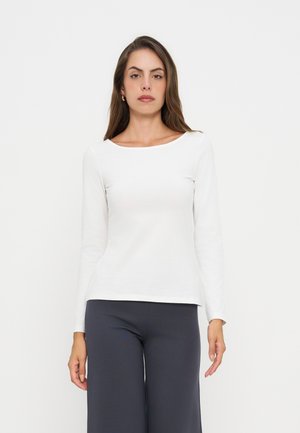 Femme aux longs cheveux bruns portant un haut blanc à manches longues et un pantalon sombre, debout devant un fond clair uni.