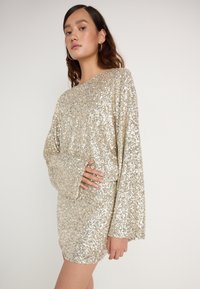 Zlata obleka s svetlečimi sekvencami z ohlapnim krojem, dolgimi rokavi in krožnim izrezom. Opozorljiv sijaj iz velikih, odsevnih sekvenc.