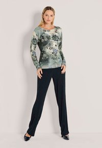 Vrouw in een lange mouwen bloempatronen top en losse zwarte broek, staande met handen rustend bij de heupen tegen een lichte achtergrond.