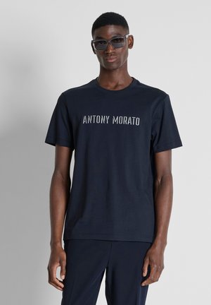 Giovane uomo di colore che indossa occhiali da sole scuri e una maglietta blu navy con "ANTONY MORATO" stampato sul davanti, in piedi davanti a uno sfondo semplice.