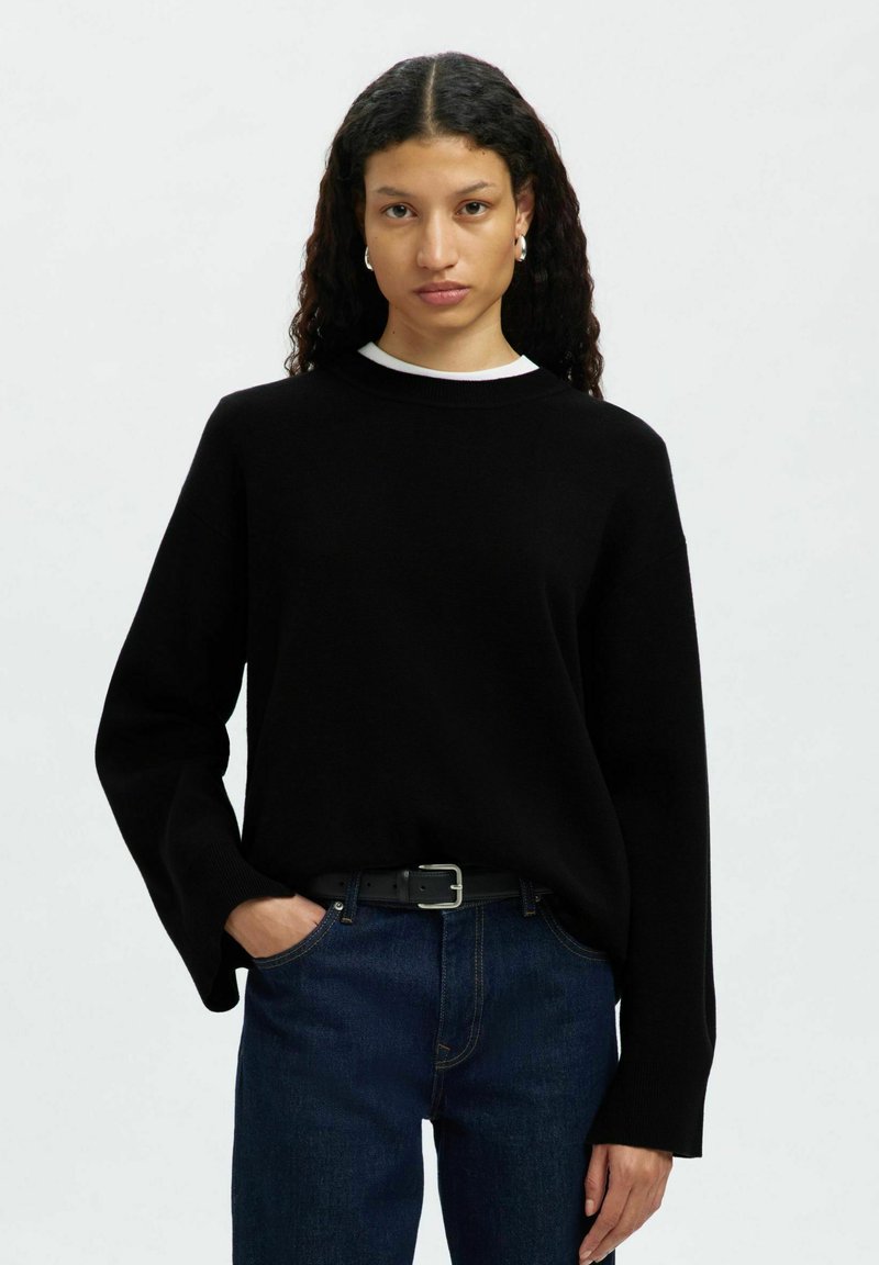 Svart stickad sweatshirt med rund halsringning, neddragna axlar och långa ärmar, i kombination med mörkblå denimjeans och ett svart bälte.