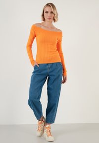 Pulover ribbed, portocalie, cu umerii goi, asociat cu pantaloni lejere din denim albastru și adidași albi cu șireturi portocalii, pe un fundal neutral.