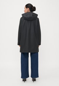 Cappotto impermeabile nero con cappuccio, dotato di maniche lunghe e taglio dritto. Indossato con jeans blu a gamba larga e scarpe con tacco.