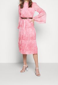 Robe rose à motifs avec manches longues évasées, design à volants, cintrée à la taille avec une ceinture marron. Associée à des sandales blanches à talons blocs.