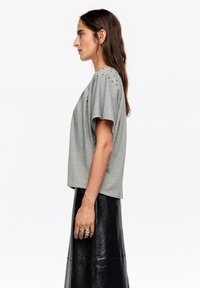 Szara koszulka z krótkimi, szerokimi rękawami i zdobieniem dziurek na ramionach, zestawiona z czarną skórzaną spódnicą. Gładka faktura materiału.