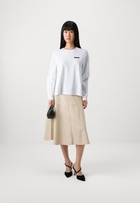 White long-sleeve shirt with a small black logo, paired with a beige flared skirt with seam details. Black handbag and heels complete the look.