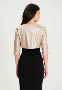 Cropped shiny beige leather jacket with three-quarter sleeves, worn over a black fitted dress, featuring a smooth texture and minimal seams.