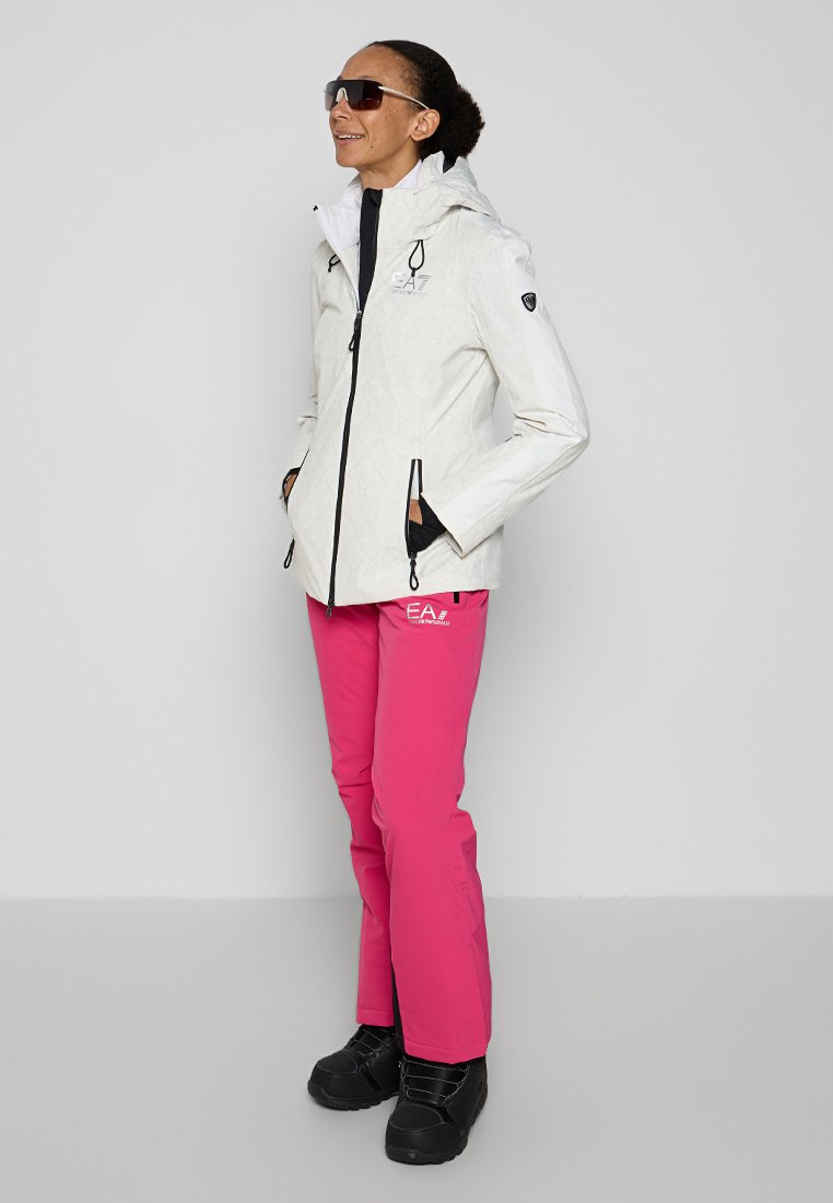 Weiße wasserdichte Jacke mit gestepptem Muster und schwarzen Akzenten, kombiniert mit leuchtend rosa Schneehosen. Schwarze Winterstiefel runden das Outfit ab.