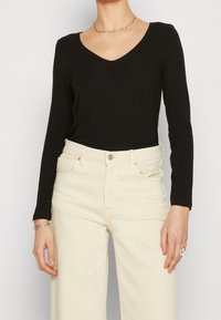Top nero a maniche lunghe con scollo a V, abbinato a jeans beige a vita alta e a gamba larga; tessuto morbido e silhouette aderente.