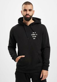 Czarny hoodie wykonany z dzianiny bawełnianej z kangurkową kieszenią, kapturem ze sznurkiem oraz białym nadrukiem "ANTI CORONA CLUB DEF" z przodu.