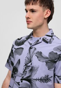 Koszula z krótkim rękawem w jasnym lawendowym kolorze z czarnym abstrakcyjnym wzorem. Posiada kołnierzyk, guziki i luźny krój. Gładka tekstura.
