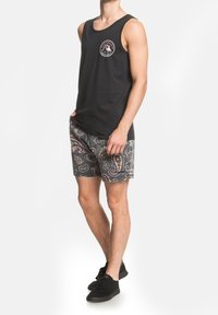 Canotta nera con un logo circolare, abbinata a pantaloni corti con motivi a paisley in tonalità di rosa e verde. Scarpe nere.