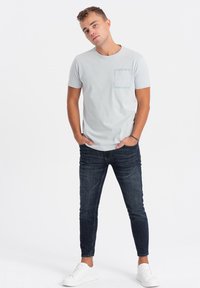 T-shirt grigio chiaro a maniche corte con taschino sul petto, abbinato a jeans slim-fit blu scuro e sneakers bianche. Design semplice e casual.
