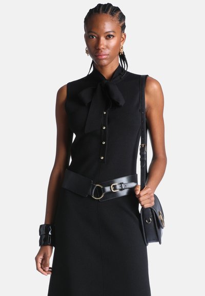Femme portant une robe noire sans manches avec un nœud au col, des boutons en perles, une large ceinture noire, un sac à main noir, des boucles d'oreilles dorées et un bracelet noir sur fond blanc.