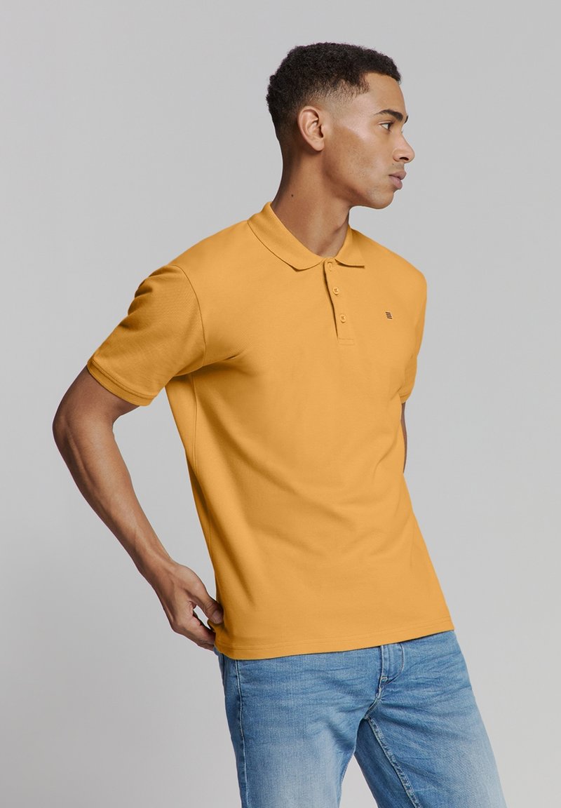 Polo shirt in mosterdgeel, gemaakt van textuurstof met een kraag en een drieknoopssluiting. Gedragen met lichtblauwe jeans.