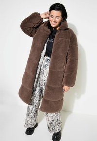 Cappotto lungo in pile marrone con una vestibilità ampia, caratterizzato da una chiusura con zip e maniche larghe, abbinato a pantaloni con stampa serpente a motivi.